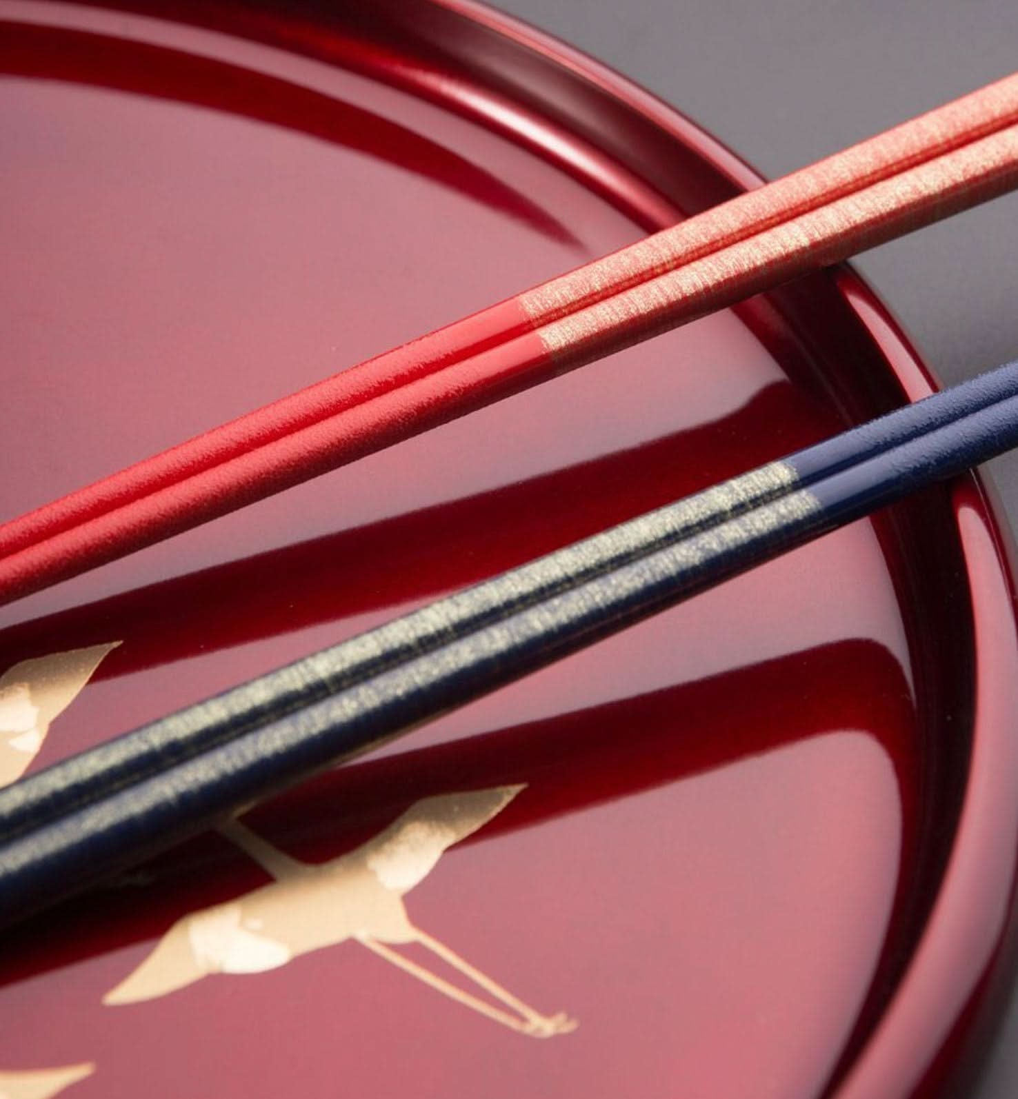 Red and navy Japanese handmade wooden chopsticks with gold accents on a red lacquered tray with crane design.